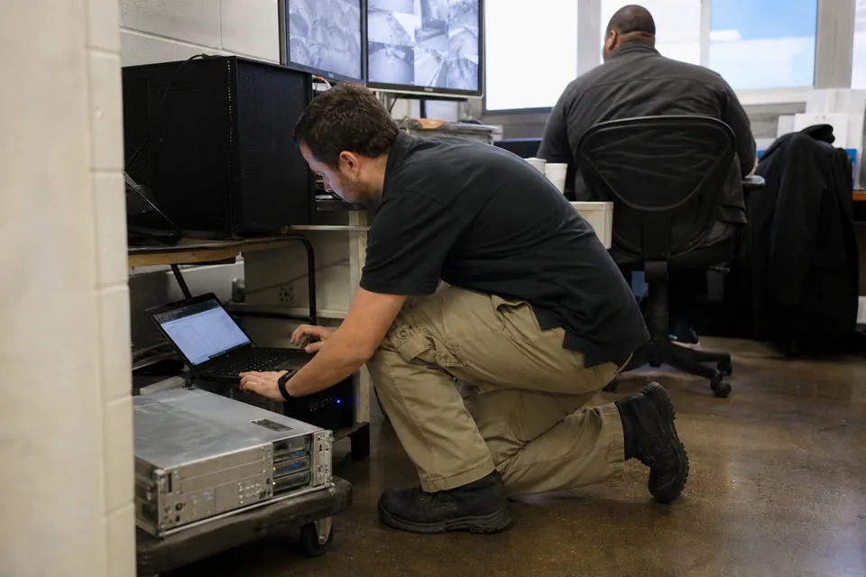IT technician beside an aging video recording workstation in a building control room, with a replacement server on a cart